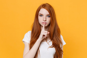 Secret young redhead woman girl in white blank empty t-shirt posing isolated on yellow background. People lifestyle concept. Mock up copy space. Saying hush be quiet with finger on lips shhh gesture.