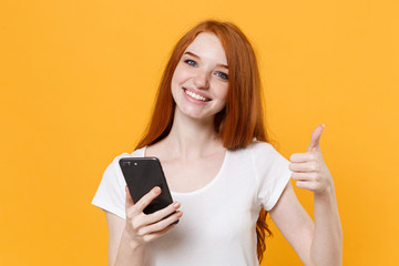 Smiling young redhead girl in white blank empty t-shirt posing isolated on yellow wall background studio portrait. People lifestyle concept. Mock up copy space. Using mobile phone, showing thumb up.