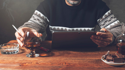 A man is sitting at a table in a tablet and Smoking a cigarette, holding a glass of cognac in his...