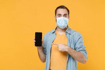 Young man in sterile face mask isolated on yellow background. Epidemic pandemic coronavirus 2019-ncov sars covid-19 flu virus concept. Pointing index finger on mobile phone with blank empty screen.