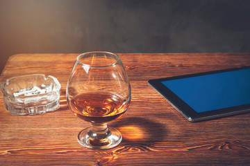On a wooden table is a tablet with a blue screen, a glass of cognac and a saucer of chocolate