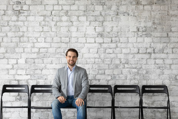 Portrait of smiling Caucasian male job candidate sit on chair in hallway wait for interview in...