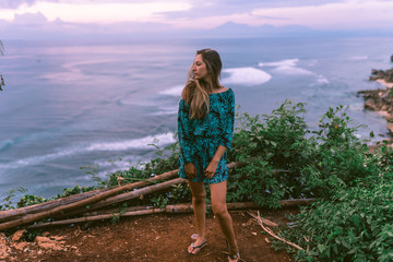 Naklejka premium A beautiful girl stands on a cliff overlooking the waves of the blue ocean. Tropical paradise and exotic nature on the island of Bali. Model with different emotions of happiness and joy.