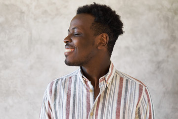 Fashionable cheerful young dark skinned man with stylish Afro haircut and bristle posing isolated in striped shirt turning head to one side, smiling broadly, keeping eyes closed, having carefree look