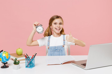 Smiling little kid schoolgirl 12-13 years old sit study at desk with laptop isolated on pink background. School distance education at home during quarantine concept. Hold alarm clock showing thumb up.