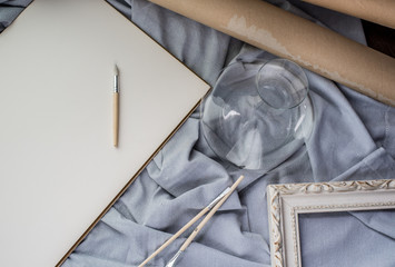 View of a creative space: brushes, vintage frame and blank paper on blue fabric.