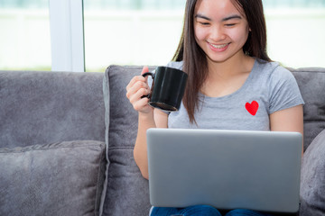 Asian woman holding a coffee cup smile and looking during a work, Working remotely with a laptop on the sofa in the house, Cute girl teens happy use internet computer to communication from home
