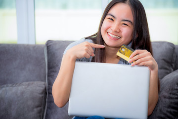 Asian young woman holding SME credit card use a laptop computer buy products payment shopping online, Cute teenage girl smile pointing finger at credit card happy lifestyle through credit card payment
