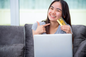 Asian young woman holding SME credit card use a laptop computer buy products payment shopping online, Cute teenage girl smile pointing finger at credit card happy lifestyle through credit card payment