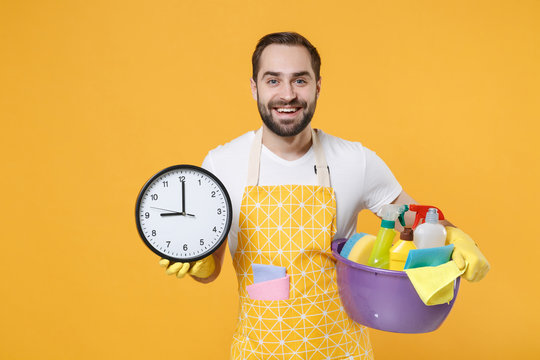 Smiling young man househusband in apron rubber gloves hold basin with detergent bottles washing cleansers doing housework isolated on yellow background in studio. Housekeeping concept. Hold clock.