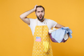 Preoccupied shocked young man househusband in apron hold basket with clean clothes doing housework...