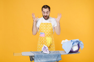 Shocked young man househusband in apron ironing clean clothes on board while doing housework...