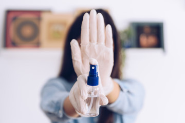 Obraz na płótnie Canvas Coronavirus concept. Girl in her home wear gloves and show as an antiseptic spray, says stop with her hand to coronavirus. Global call to stay home.