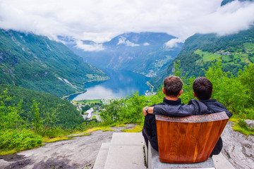 Male tourists sitting on the Queen Sony Chair at the Flydalsjuvet Viewpoint. The Geiranger village...