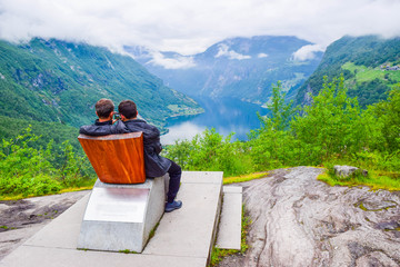 Fototapeta na wymiar Male tourists sitting on the Queen Sony Chair at the Flydalsjuvet Viewpoint. The Geiranger village and Geirangerfjord landscape. Norway.