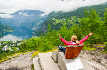 Fototapeta na wymiar Female tourist sitting on the Queen Sony Chair at the Flydalsjuvet Viewpoint. The Geiranger village and Geirangerfjord landscape. Norway.