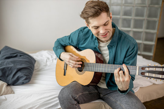 young caucasian guy play acoustic guitar at home, man keen on music, perform music. handsome male in casual wear sit on bed with guitar. indoors