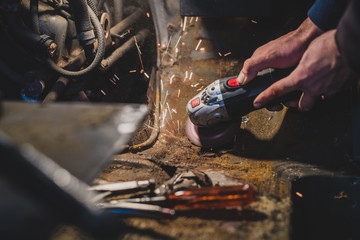 Bare hands of a man using an angle grinder to cut off the excess rusty metal from a car body in the attempt to restore it and weld in a new piece. Sparks are flying around, tools and grinding plates l