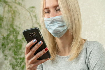 Blonde girl in a medical mask looks at a black mobile phone in her hand. Quarantine during the coronavirus pandemic on November 19.
