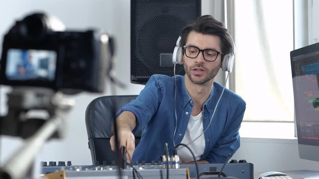 Young man in sound recording studio. Video blogger explain on camera how to use mixing console and work with it. Listening music through headphones.