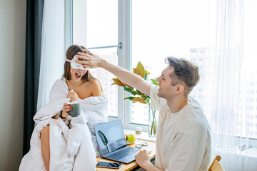 Fototapeta premium adorable caucasian lady is next to her husband while he works in the morning. man use laptop, he is at freelance work. cute wife wrapped in blanket miss him, wait for his free time