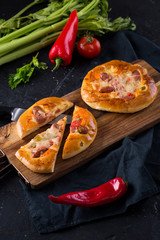 Home made small pizzas with tomatoes and cheese on wooden cutting board on dark background with wheat spikes. Healthy proper food preparation, copy space