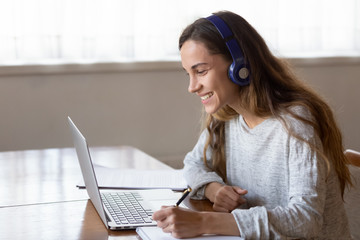 Head shot side view smiling young mixed race girl wearing wireless headphones, looking at laptop...
