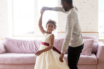 Loving african american father holding hand of adorable daughter dancing to favorite song at home. Diverse happy smiling dad teaching his young girl and twisting cute little kid in living room. - obrazy, fototapety, plakaty