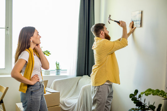young caucasian married couple hanging picture on the wall of living room, they decorate together. man use hammer. after moving into new apartment