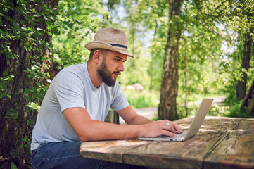 Bearded man is working and smiling with laptop at the park under the tree. Happy freelancer is sitting and using app or website on the grass. Hipster is texting email or chating at social networks