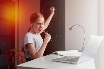 Joyful teenage girl in headphones rejoices and sings, performs school tasks at home, searches for...