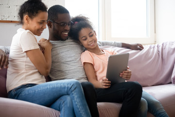 Happy african american couple looking at tablet screen with daughter. Diverse smiling family of mother, father and girl sitting on couch using mobile device and gadget together.