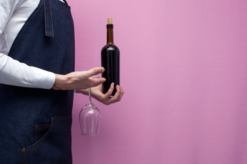 professional waiter in uniform holds a bottle of red wine and a glass on a pink background, close-up