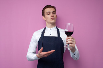 young sommelier guy in a white shirt and apron holds a glass of red wine and appreciates the taste, an expert tastes wine on a colored background