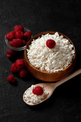 fresh homemade cottage cheese in a wooden bowl with raspberries on a dark background, healthy food on a black table