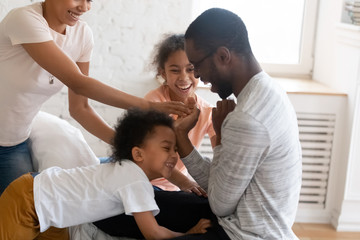 Happy african american family tickle children and having good time on bed. Young diverse smiling husband and wife with cute daughter and son sitting in bedroom at home playing with kids.