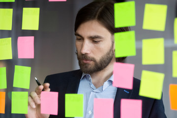 Pensive middle-aged Caucasian businessman do creative thinking brainstorm on company financial business project in office, thoughtful male employee write ideas on colorful sticky notes on glass wall
