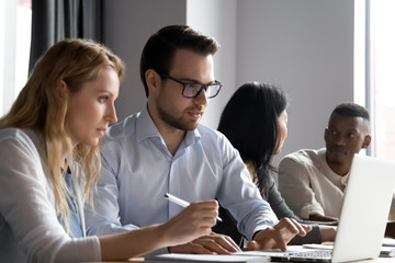 Concentrated diverse colleagues sit at desk brainstorm work on laptop together, focused multiracial...