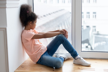 Pensive african american young girl sitting alone on windowsill in living room at home. Young...