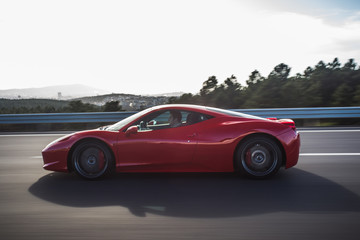 Naklejka na ściany i meble Red luxury model sport car profile view on the highway across green hills