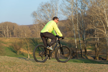 Obraz premium Cyclist in pants and green jacket on a modern carbon hardtail bike with an air suspension fork. The guy on the top of the hill rides a bike.