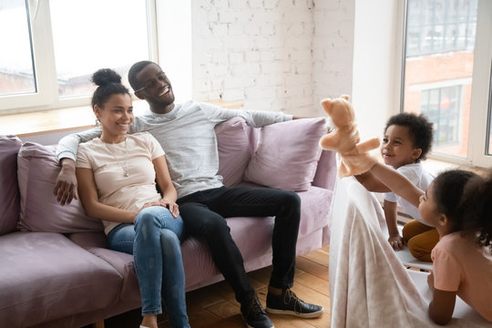 Happy african american couple watching cute siblings puppet show. Smiling young father and mother sitting together on sofa at home, brother and sister play muppet toys do theatre performance.