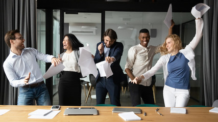 Overjoyed diverse businesspeople have fun in boardroom celebrate successful project together, happy excited multiracial colleagues dance laugh with paperwork in office, Friday celebration concept