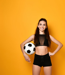 Brunette model dressed in black top and shorts. Smiling, put hand on hip, holding soccer ball, posing on orange studio background