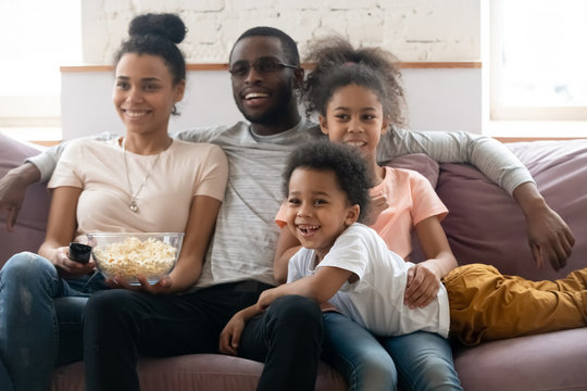 Happy african american family with kids watching funny tv show or movie eating popcorn snack. Happy diverse dad holding remote controller, mom hugging cute siblings.