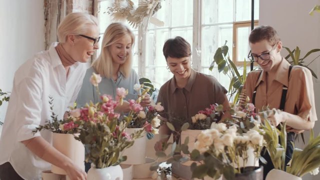 Team of mixed aged cheerful male and female florists chatting and laughing while arranging flower compositions on masterclass in workshop