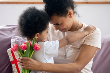 Close up african american mother holding flowers, bouquet and gift embracing little son,...