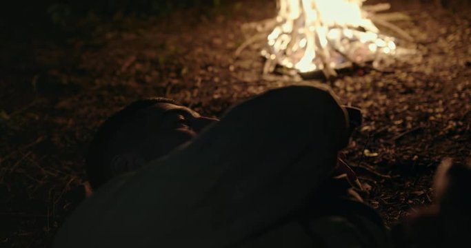Soldier sleeping on the ground near a blazing campfire at night in a close up view as he turns restlessly under his blanket