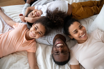 Happy african american parents with little children lying on bed top view, looking at camera,...