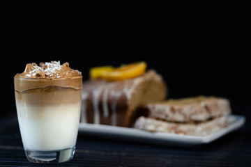 Iced Dalgona or whipped coffee on a glass and a orange cake behind. Black background